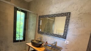 Un baño moderno con tocador de madera, dos lavamanos sobre encimera, un espejo grande y una ducha con mampara de vidrio.