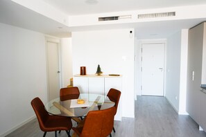 A modern dining area with a glass table and four brown chairs, a white door, and a small shelf with a plant and a vase. - Pine Hills - New Apartment Fuengirola (Fuengirola)