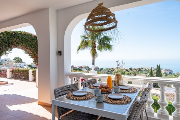 A dining table with plates, cups, and a bottle, set against a scenic view of the sea and hills. - Villa Sabine - Spectacular Sea View Villa in Capel (Benalmádena)