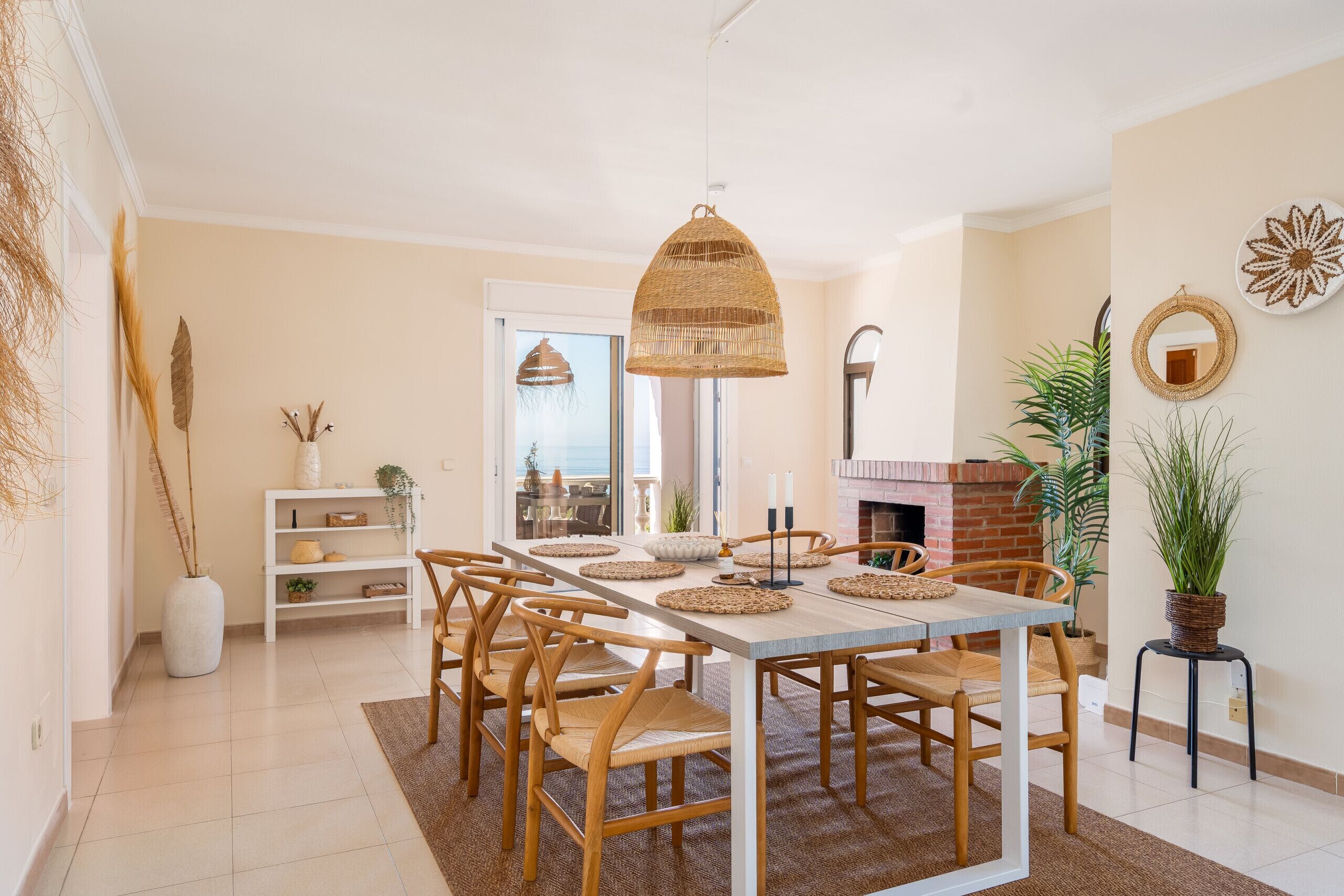 A dining area with a large table, chairs, a hanging light fixture, a brick fireplace, and a potted plant.