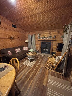 A wooden cabin interior with a sofa, a round coffee table, a fireplace, and a rocking chair.