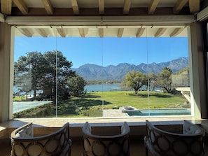 A view from a room with a large window overlooking a lake, mountains, and a well-maintained lawn with a pool and seating area.