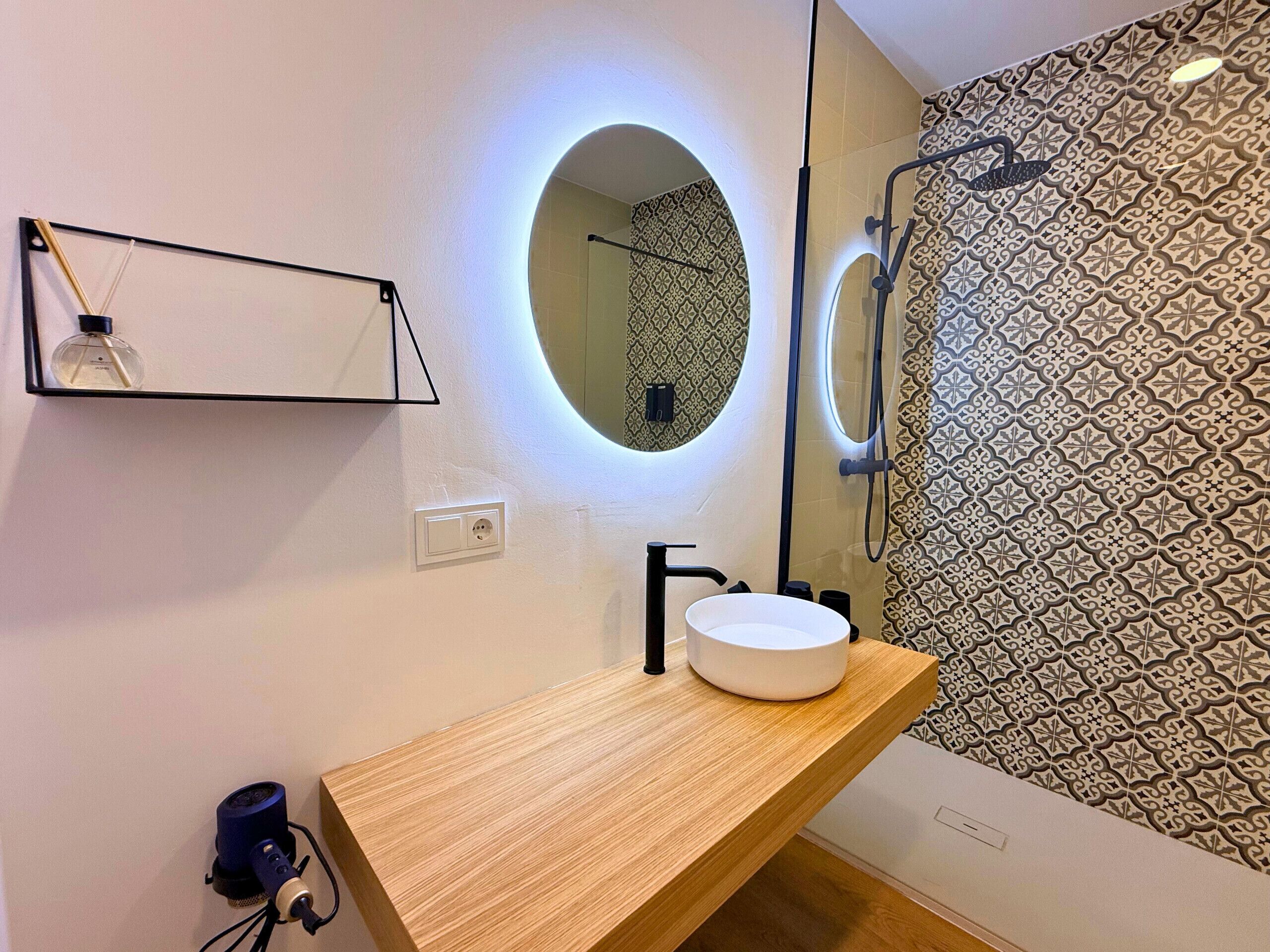 A bathroom with a wooden vanity, a round mirror with underglow lighting, a black faucet, and patterned shower walls.