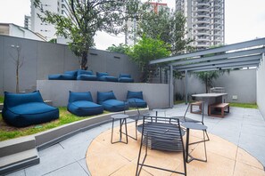 Uma área de estar moderna ao ar livre, com cadeiras estofadas em azul e uma mesa de metal, cercada por um muro baixo e vegetação.