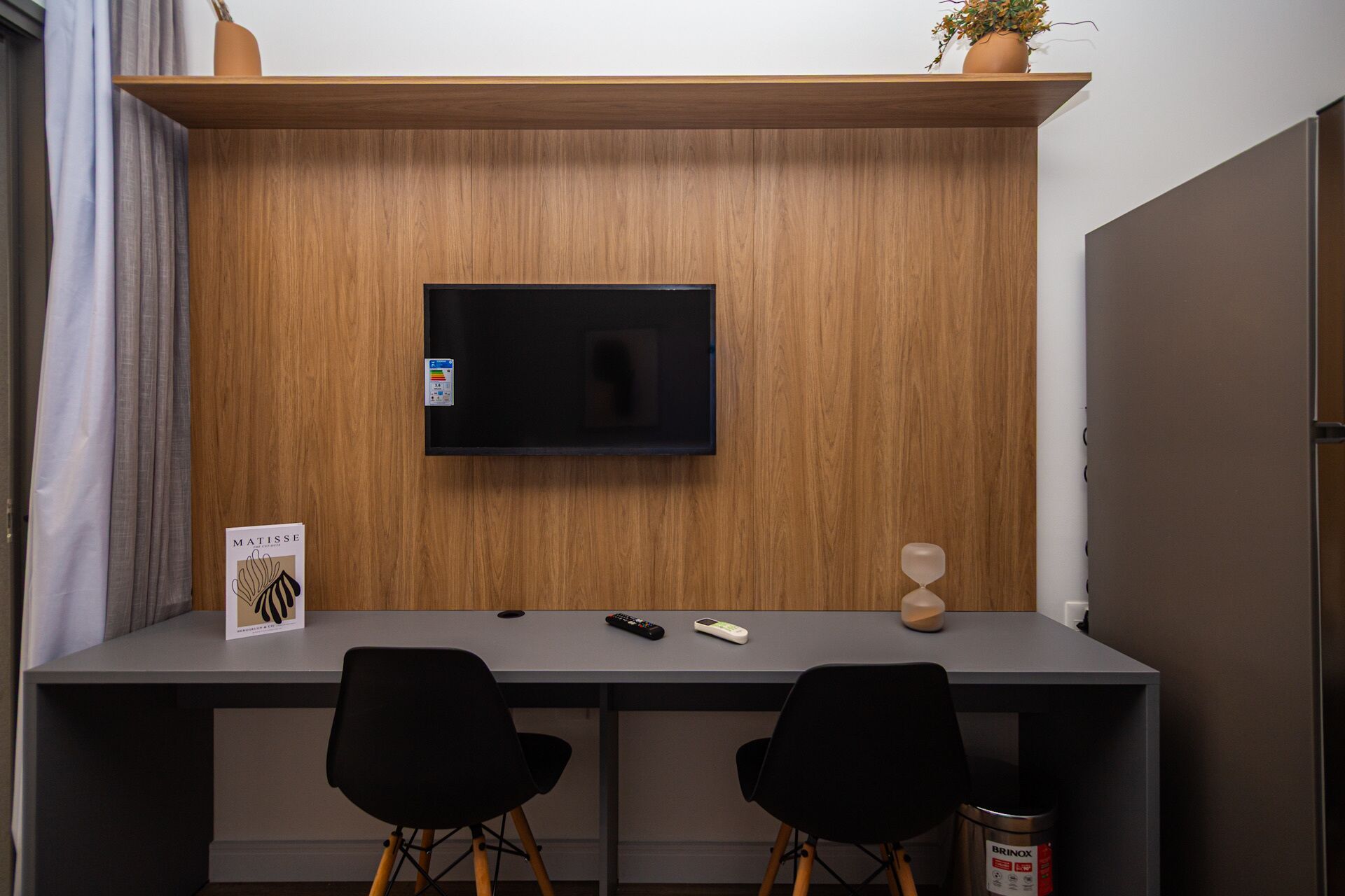Ein modernes Zimmer mit Holzwand, einem montierten Fernseher, zwei schwarzen Stühlen, einer Fernbedienung und einem kleinen Topfpflanzen.