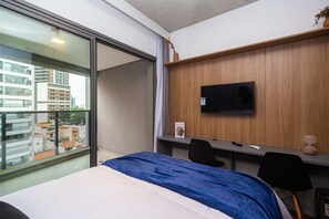 A hotel room with a bed, a desk with a chair, a TV mounted on the wall, and a balcony with a view of buildings. - Studio no coração da Vila Madalena (São Paulo)
