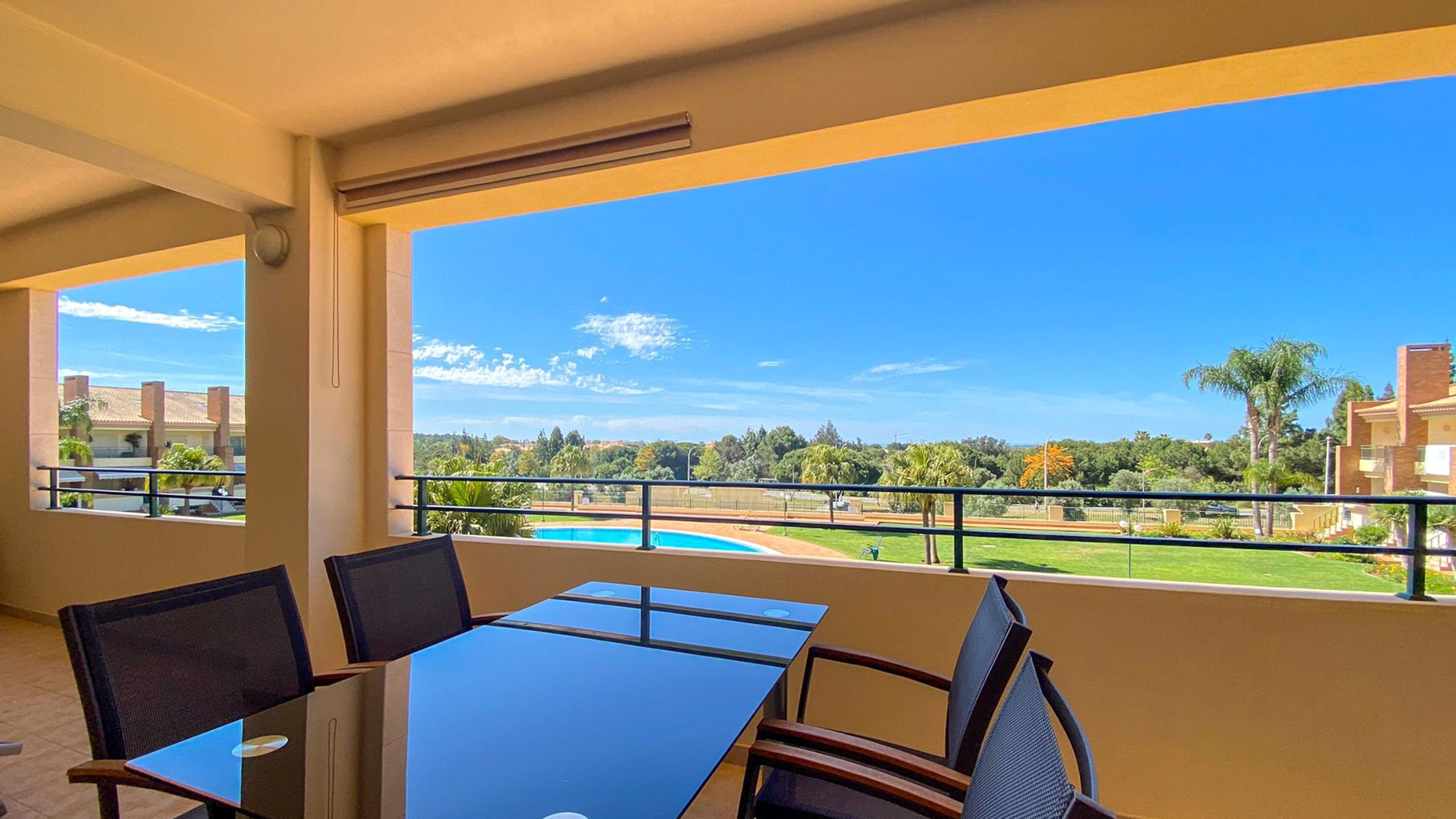 A balcony with a glass table and chairs, overlooking a swimming pool and greenery.