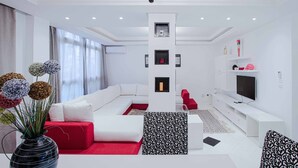 Una sala de estar moderna con un sofá blanco, almohadones rojos decorativos, un reposapiés blanco y negro estampado, una araña y un televisor de pantalla plana sobre un mueble blanco.