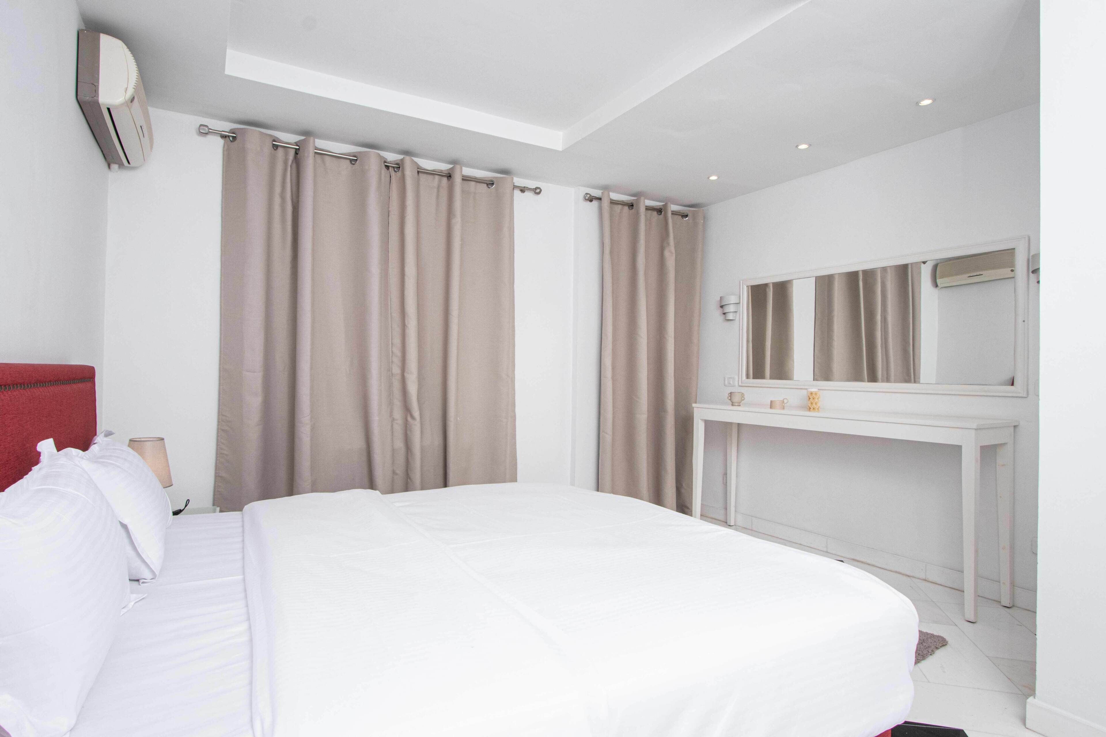A hotel room with a large bed, a white bedside table with a mirror, and beige curtains.