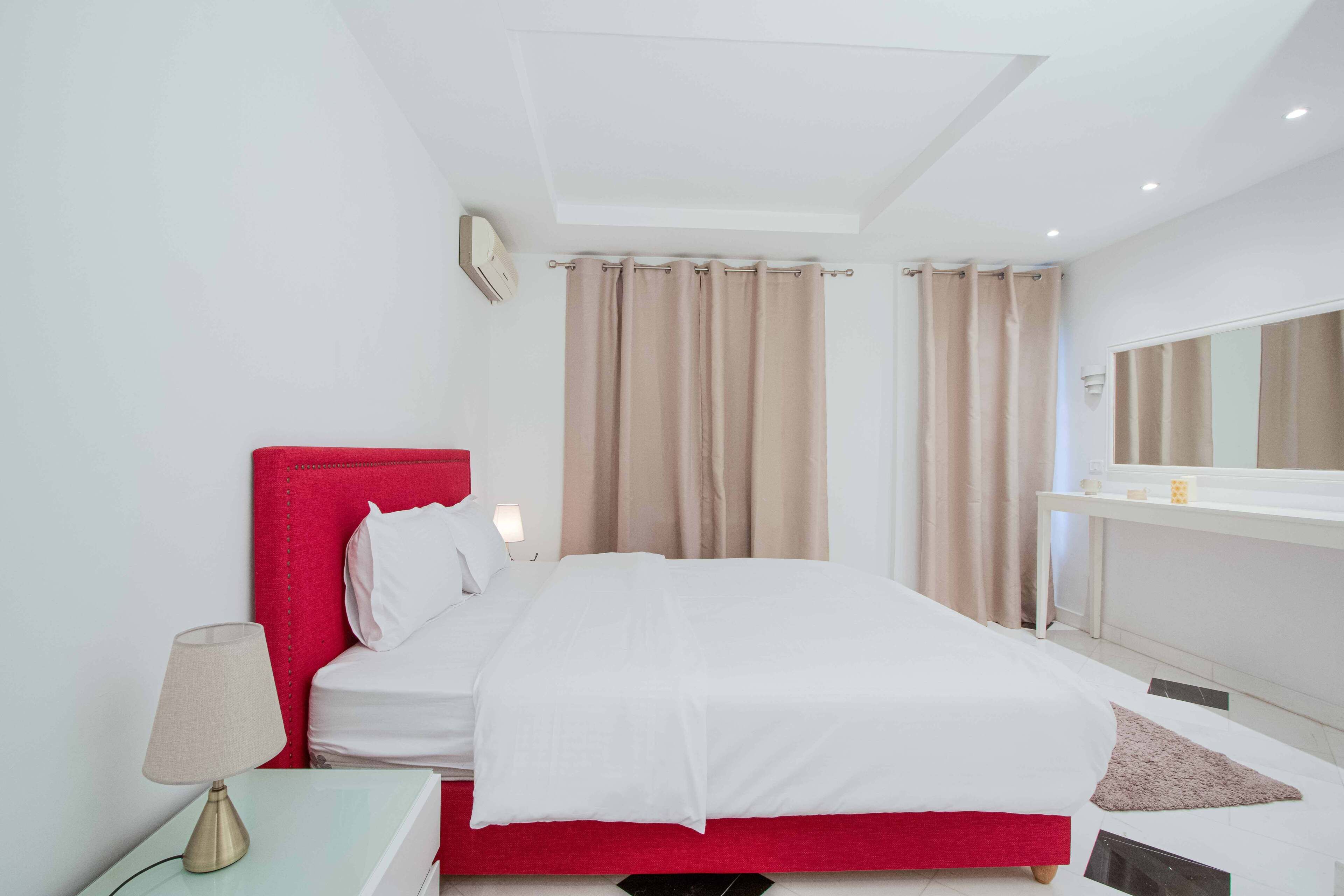 A hotel room with a red headboard, white bedding, a bedside lamp, a mirror, and beige curtains.