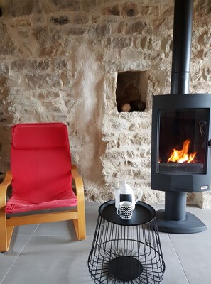 A stone wall, a red chair, a black table with a coffee pot and mug, and a wood-burning stove.