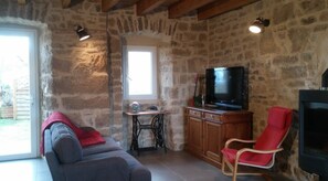 Une piÚce aux murs de pierre, avec un téléviseur, un fauteuil rouge, un canapé gris, une petite table avec des livres et une machine à coudre.