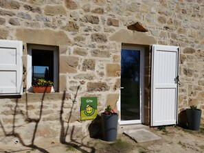Un bĂątiment en pierre avec une porte et une fenĂȘtre, une enseigne indiquant quâil sâagit dâun gĂźte, et des plantes en pot.