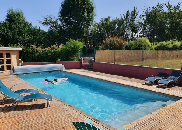 A swimming pool with a wooden deck, lounge chairs, and a small building.