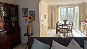 En stue med en mørkebrun sofa, et træskab, en vase med solsikker, et spisebord med stole og udsigt til det fri gennem et vindue.