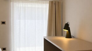 Une chambre moderne équipée d’un bureau, d’une chaise, d’une fenêtre avec des rideaux légers et d’une lampe.