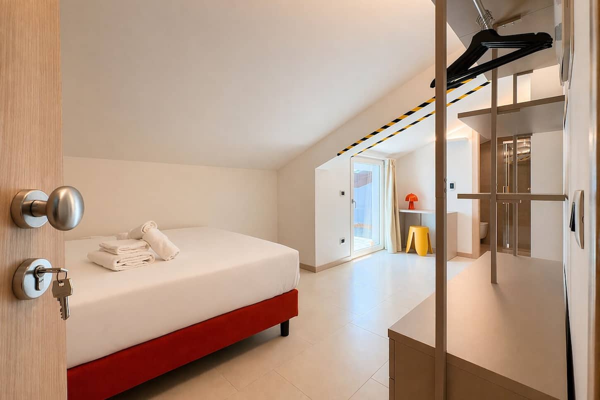 A modern hotel room with a bed, a bathroom, and a hallway leading to another room.