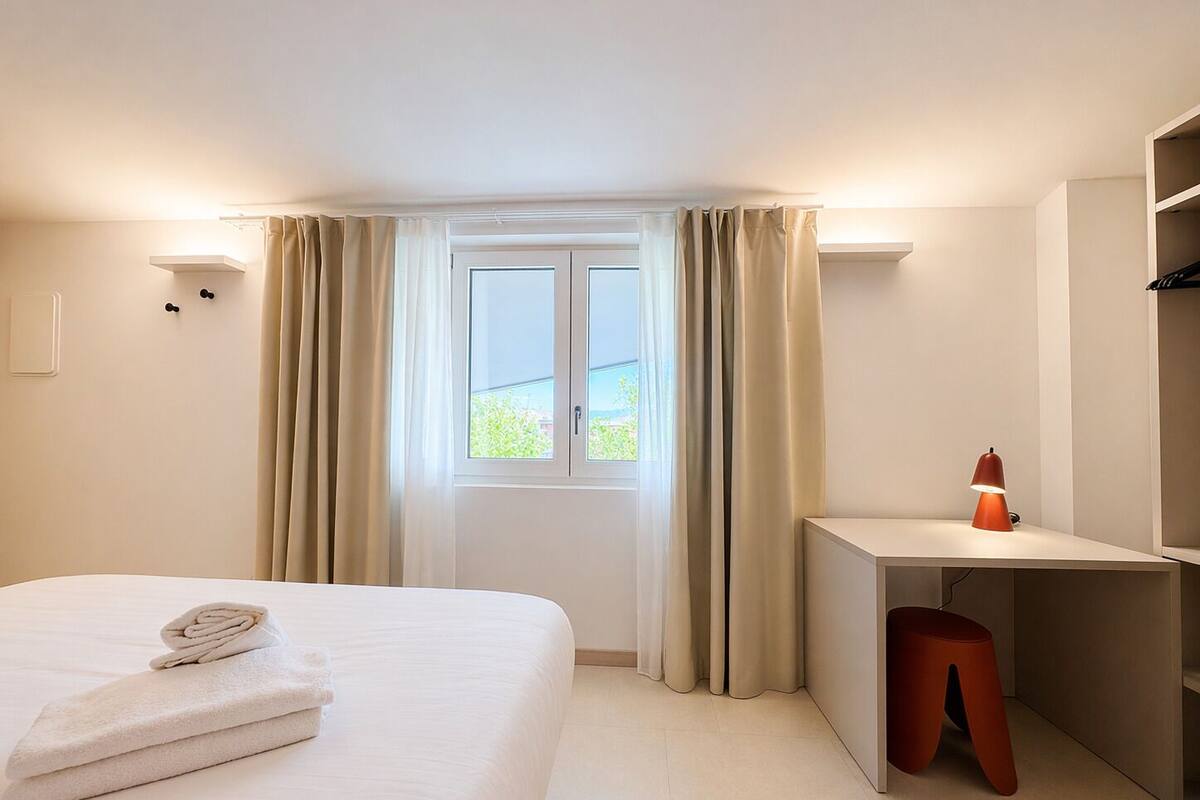 A hotel room with a bed, a desk, a chair, a lamp, and a window with curtains.