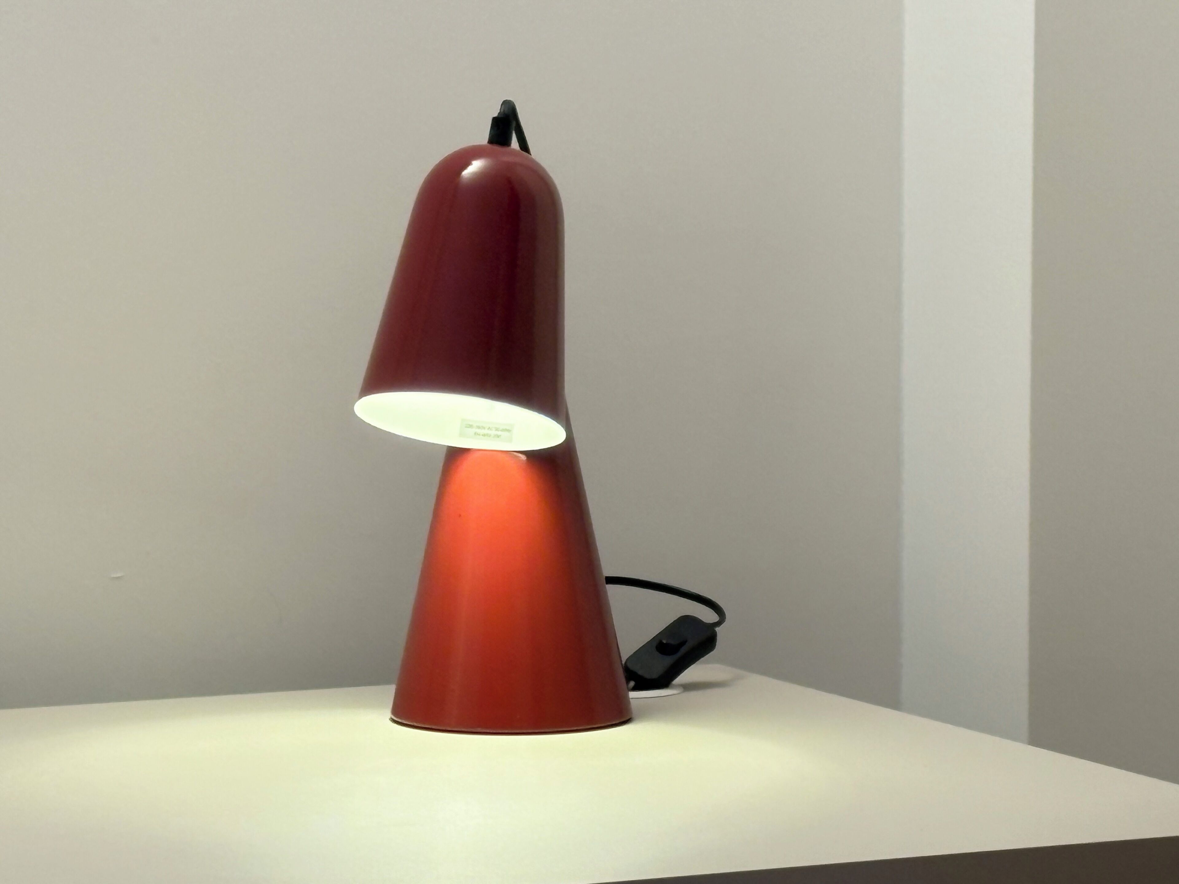 A red, conical desk lamp with a white base and a power cord plugged in.