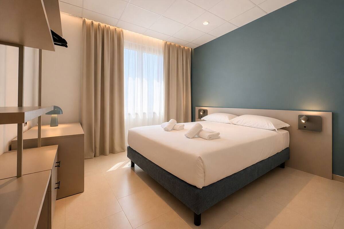 A modern hotel room with a bed, bedside tables, a desk, and a window with curtains.