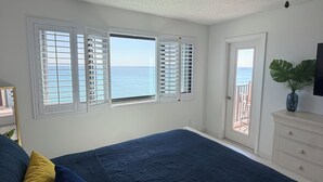 Ein Schlafzimmer mit einem großen Bett, zwei Fenstern mit Rollläden, einem Deckenventilator, einem Schminktisch und Blick auf den Ozean.