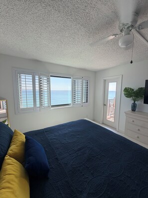 Ein Schlafzimmer mit einem großen Bett, zwei Fenstern mit Rollläden, einem Deckenventilator, einem Schminktisch und Blick auf den Ozean.