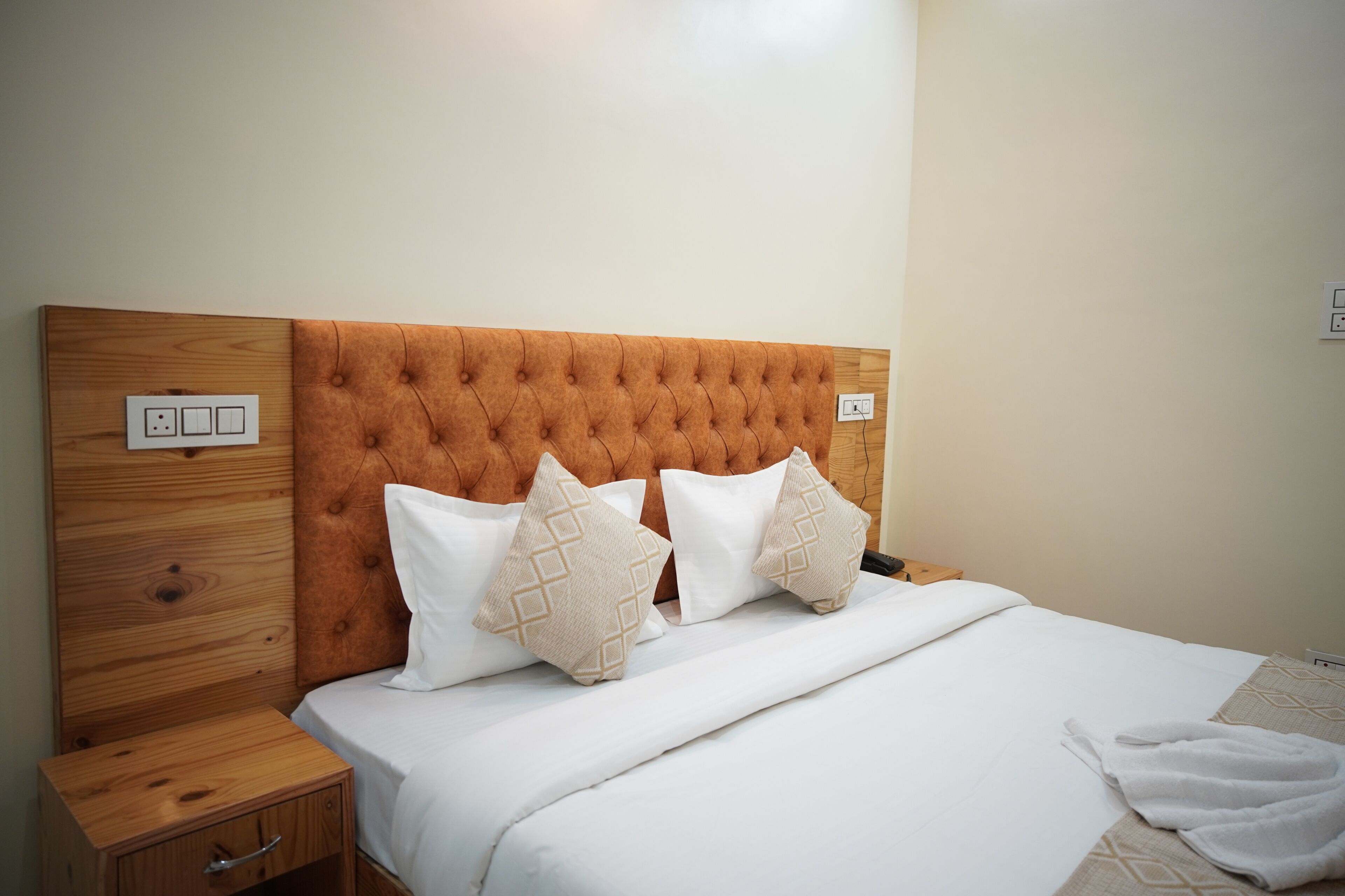 Uma cama bem arrumada com cabeceira de madeira, mesa de cabeceira com gaveta e tomada na parede.