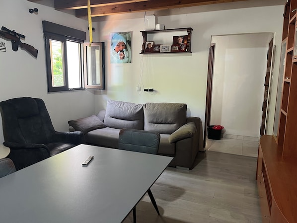 A room with a sofa, a chair, a table, a door, and a wall-mounted shelf with pictures.