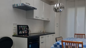 Una cucina compatta con mobili bianchi, forno a incasso, una sedia nera, un tavolo da pranzo con tovaglia a quadretti blu e una lampada a sospensione.
