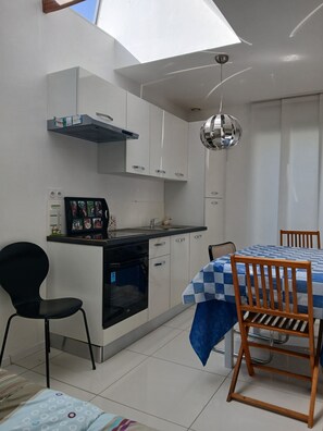 Une cuisine compacte avec des meubles blancs, un four encastré, une chaise noire, une table à manger recouverte d’une nappe à carreaux bleus et un luminaire suspendu.