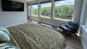 Un dormitorio con cama, una silla, ventilador de techo, televisor y ventanas amplias con vistas a árboles y un lago.
