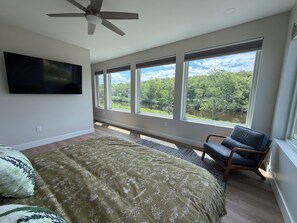 Une chambre à coucher comprenant un lit, une chaise, un ventilateur de plafond, un téléviseur et de grandes fenêtres donnant sur des arbres et un lac.