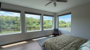 Camera da letto con un letto grande, un ventilatore a soffitto e tre finestre che danno su una foresta e un fiume.