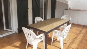 Eine Terrasse mit Holztisch und weißen Plastikstühlen, einer Schiebetür und einer kleinen Küchenzeile.
