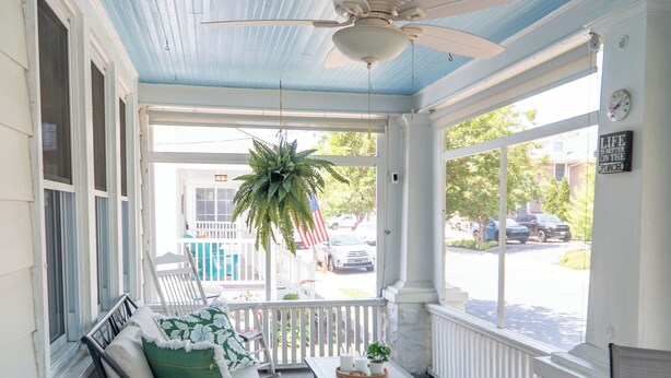Une véranda dotée d’un ventilateur de plafond, un banc avec des coussins, une petite table avec une plante, et une vue sur la rue.