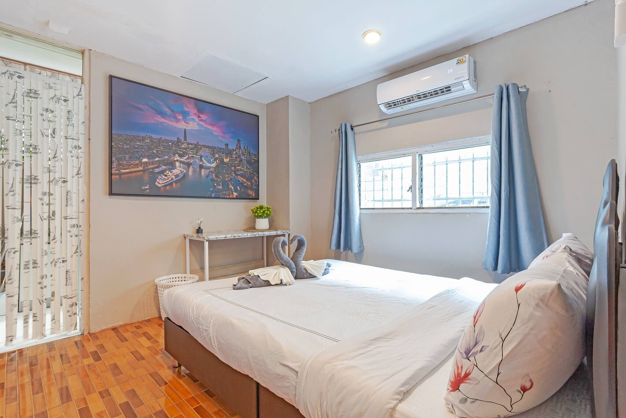 A hotel room with a bed, a window with curtains, a wall-mounted air conditioner, a cityscape painting, and a small plant on a side table.