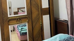 A wooden wardrobe with a mirror, a bed with a blue bedspread, and a window with curtains.