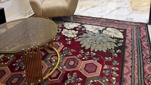 A room with a plush armchair, a dining table with chairs, a patterned carpet, and a view of the outside.