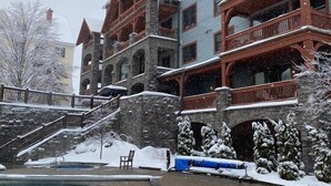 A multi-story building with a stone facade, wooden balconies, and a pool area with a brick deck. - Luxury Stratton Village Condo | Walk to Lifts, Dining & Amenities (Stratton,)