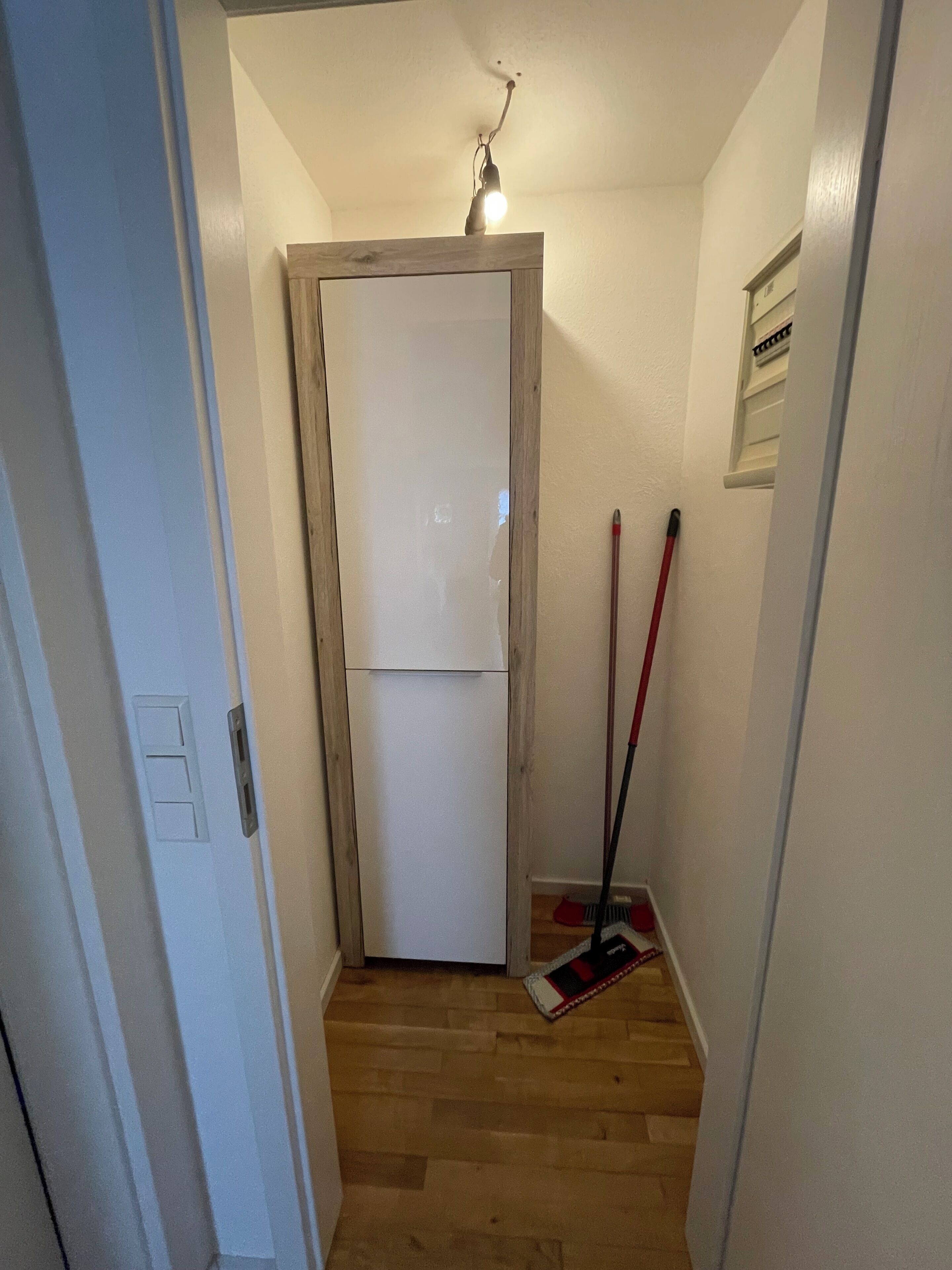 Un pasillo estrecho con una puerta de madera, una escoba y un trapeador apoyados en la pared.