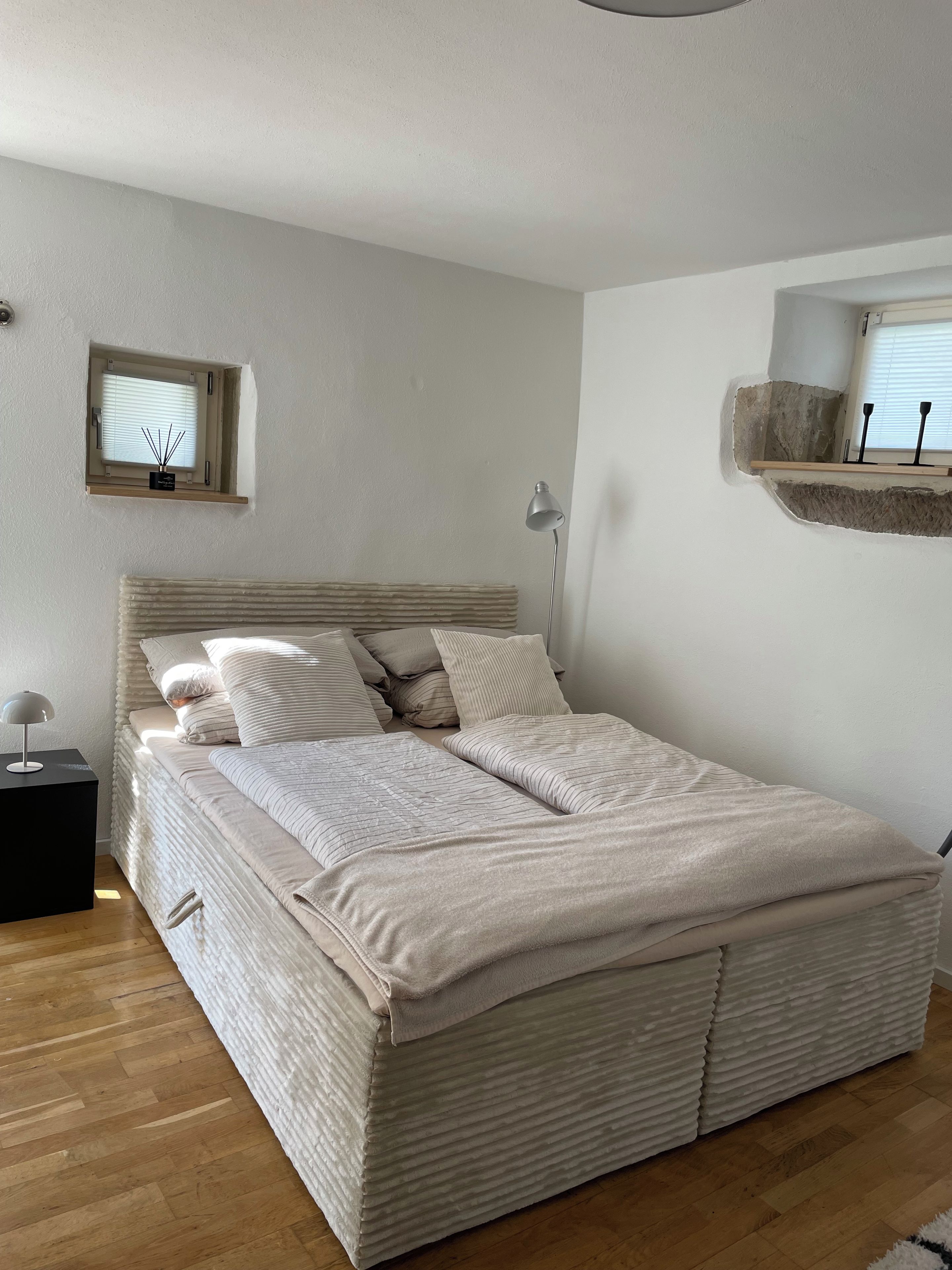 A bedroom with a bed, bedside table, lamp, window, and a shelf with decorative items.