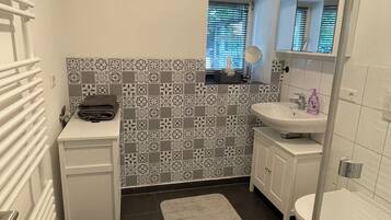 Une salle de bain avec un lavabo blanc, un miroir, des toilettes, un pare-feu décoratif et une fenêtre avec des stores.