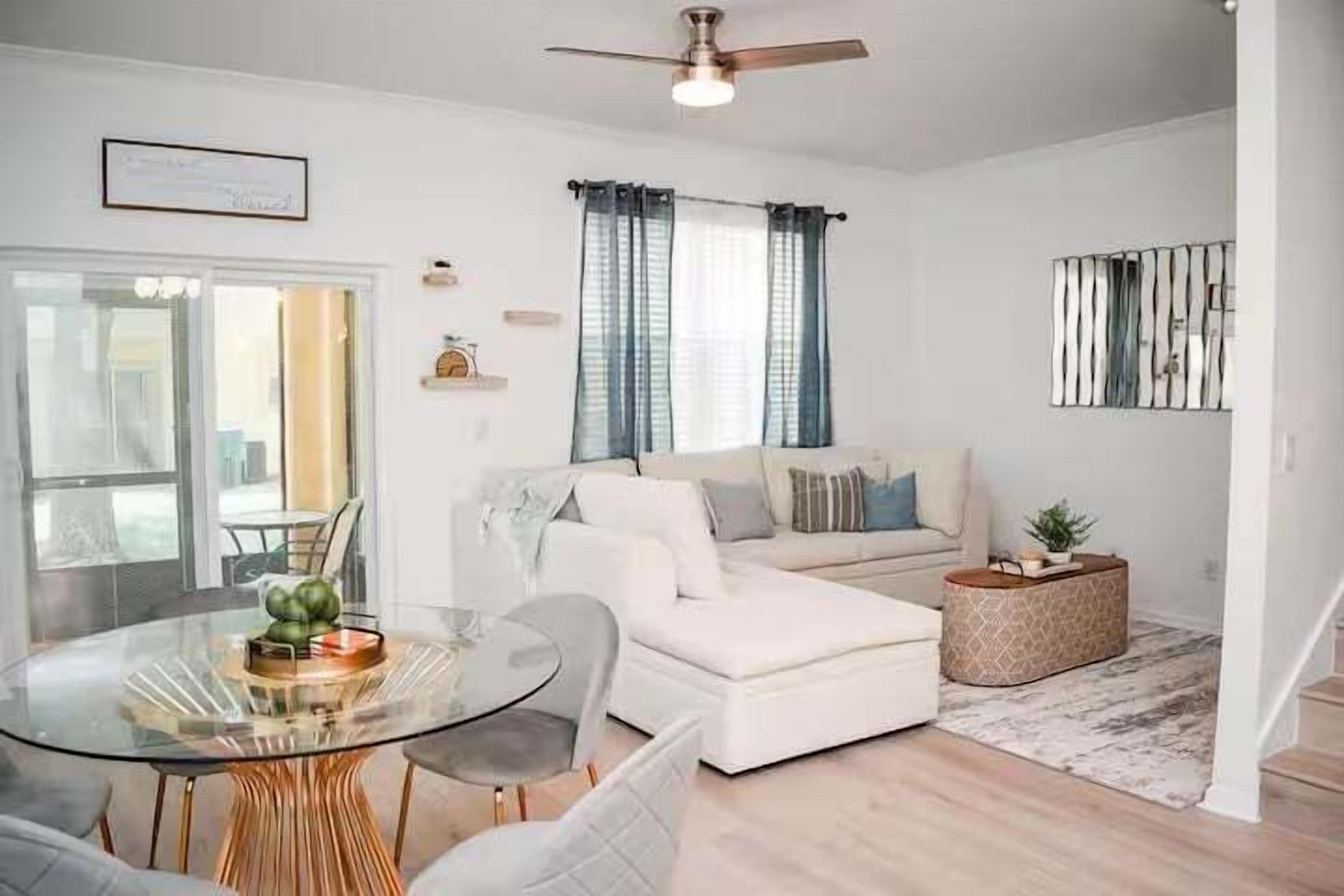 A modern living room with a glass coffee table, a white sofa, a round ottoman, and a ceiling fan.