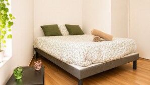 A bed with a floral bedspread, two pillows, a rolled towel, a green plant, and a small table with a vase and a shoe. - Cosy apartment in the heart of Angers historic district – Terrace & comfort – Sleeps 2 (Angers)