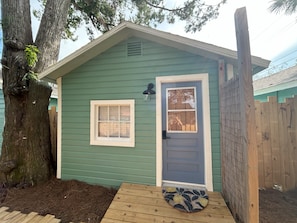 A small, single-story house with a green exterior, a blue door, a small window, and a wooden porch.