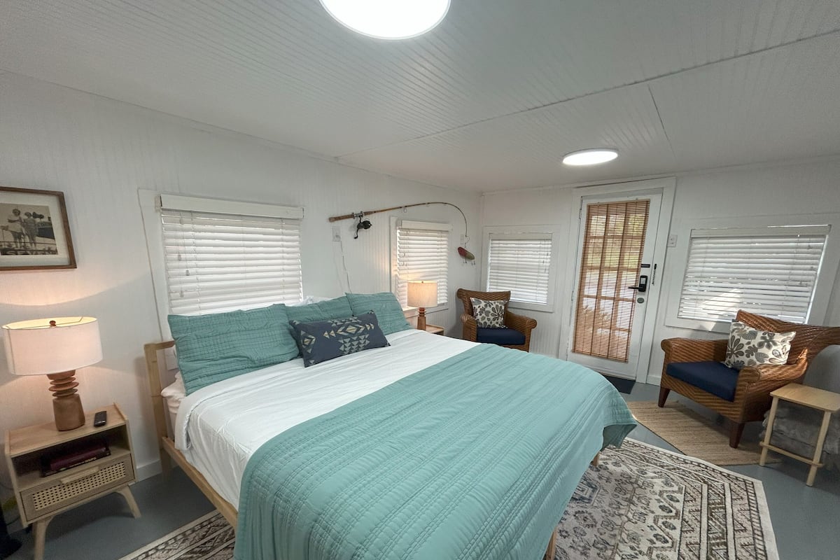 Un dormitorio con cama, una silla, una mesita de noche, una lámpara y una ventana con persianas.