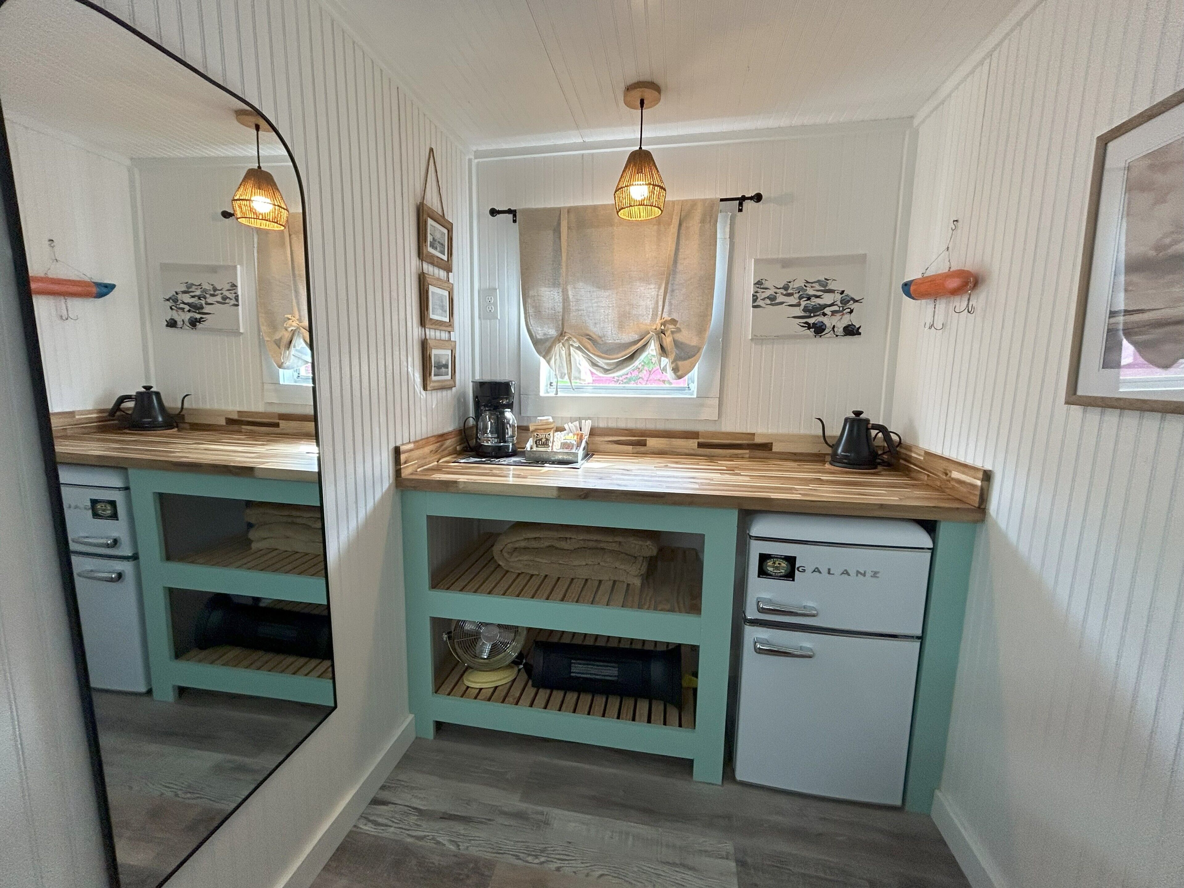 A compact kitchen with a mirrored backsplash, wooden countertops, a coffee maker, a fan, and a small refrigerator.