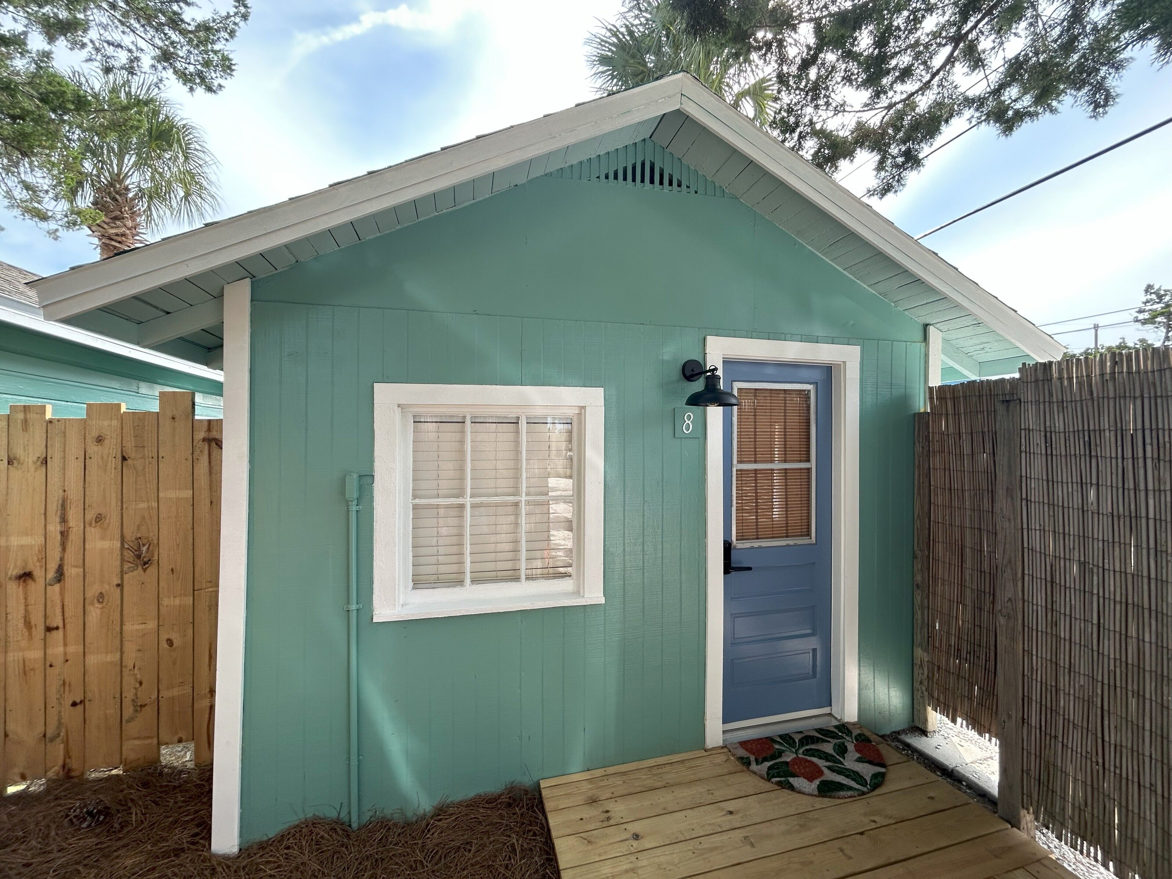 A small, single-story building with a blue door and a window, surrounded by a wooden fence and a wooden deck.