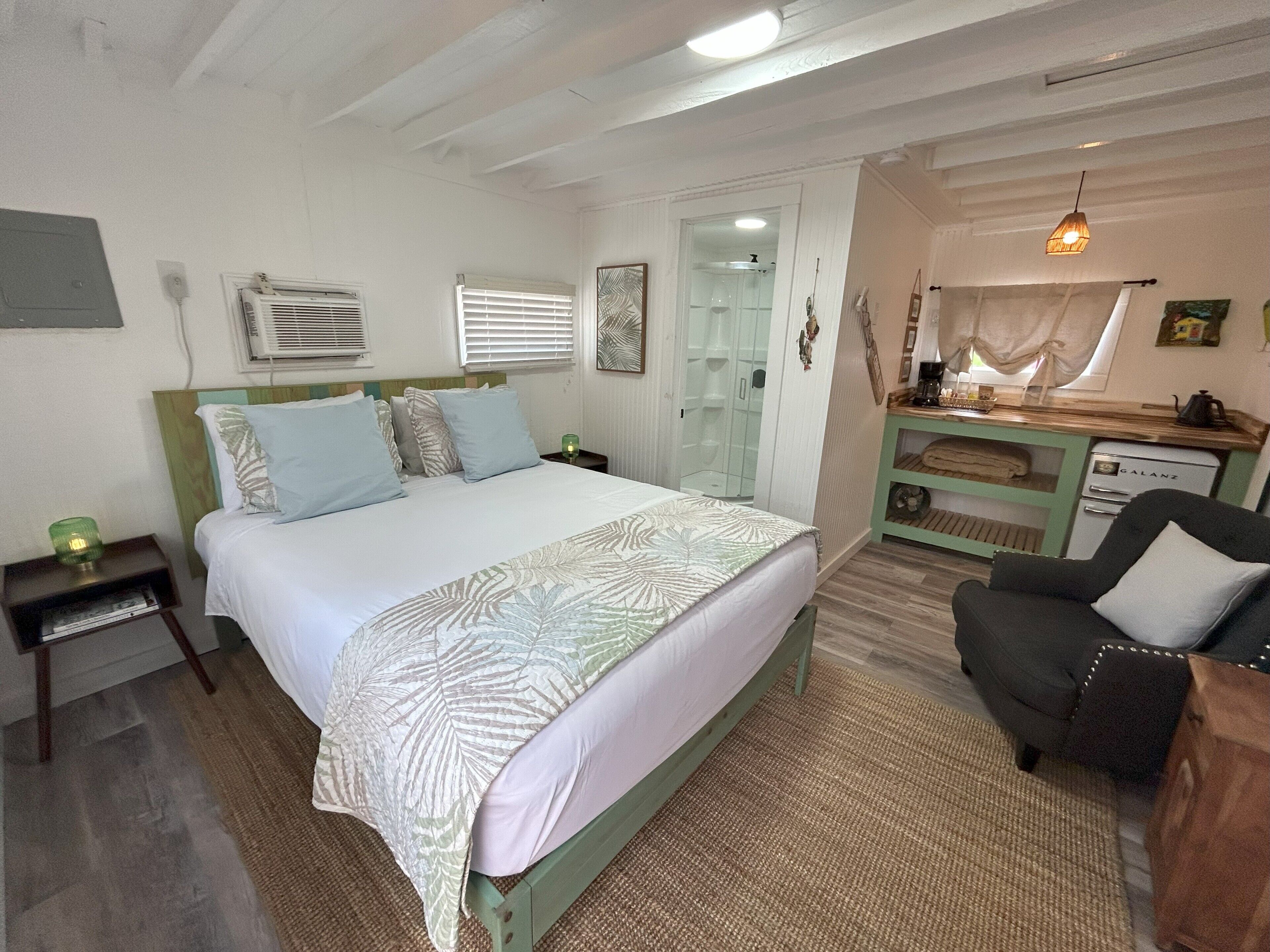 A bedroom with a bed, bedside tables, a desk, a chair, a hanging light fixture, and a shower area.