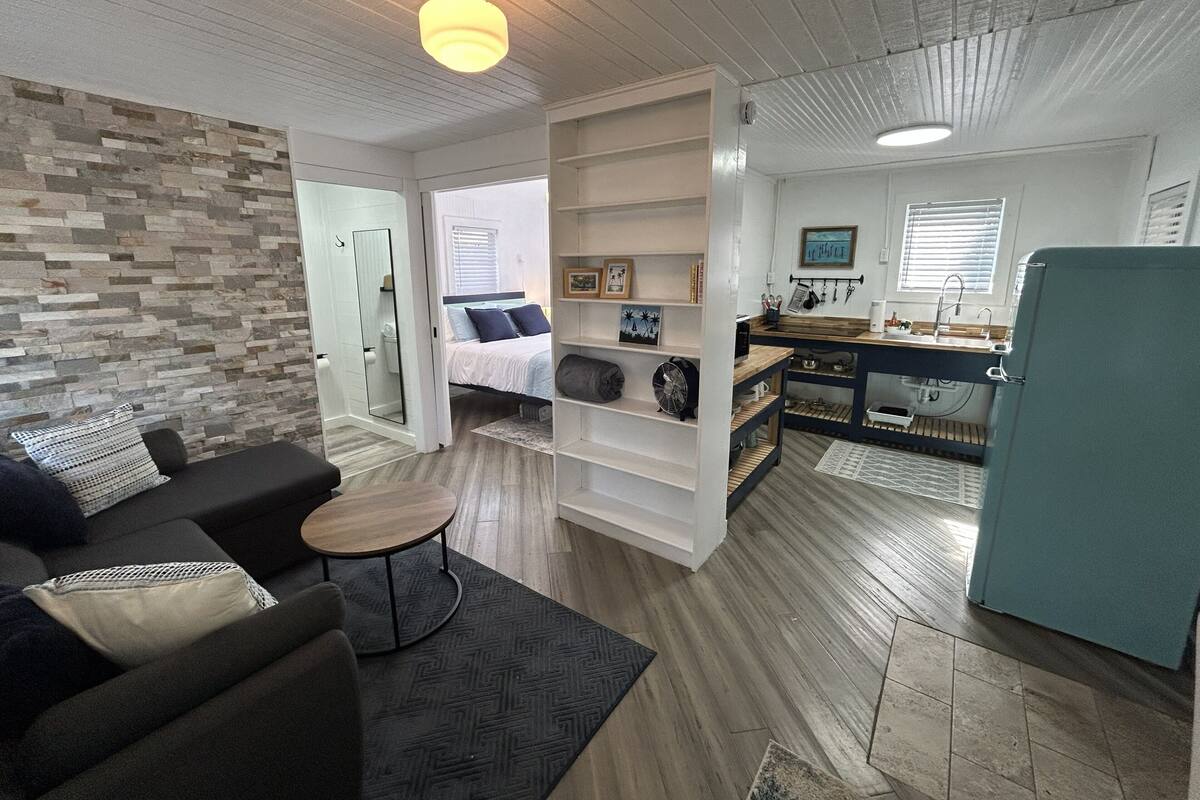 Un espacio habitable compacto con sofá, una mesita, estantería empotrada, kitchenette y baño con ducha.